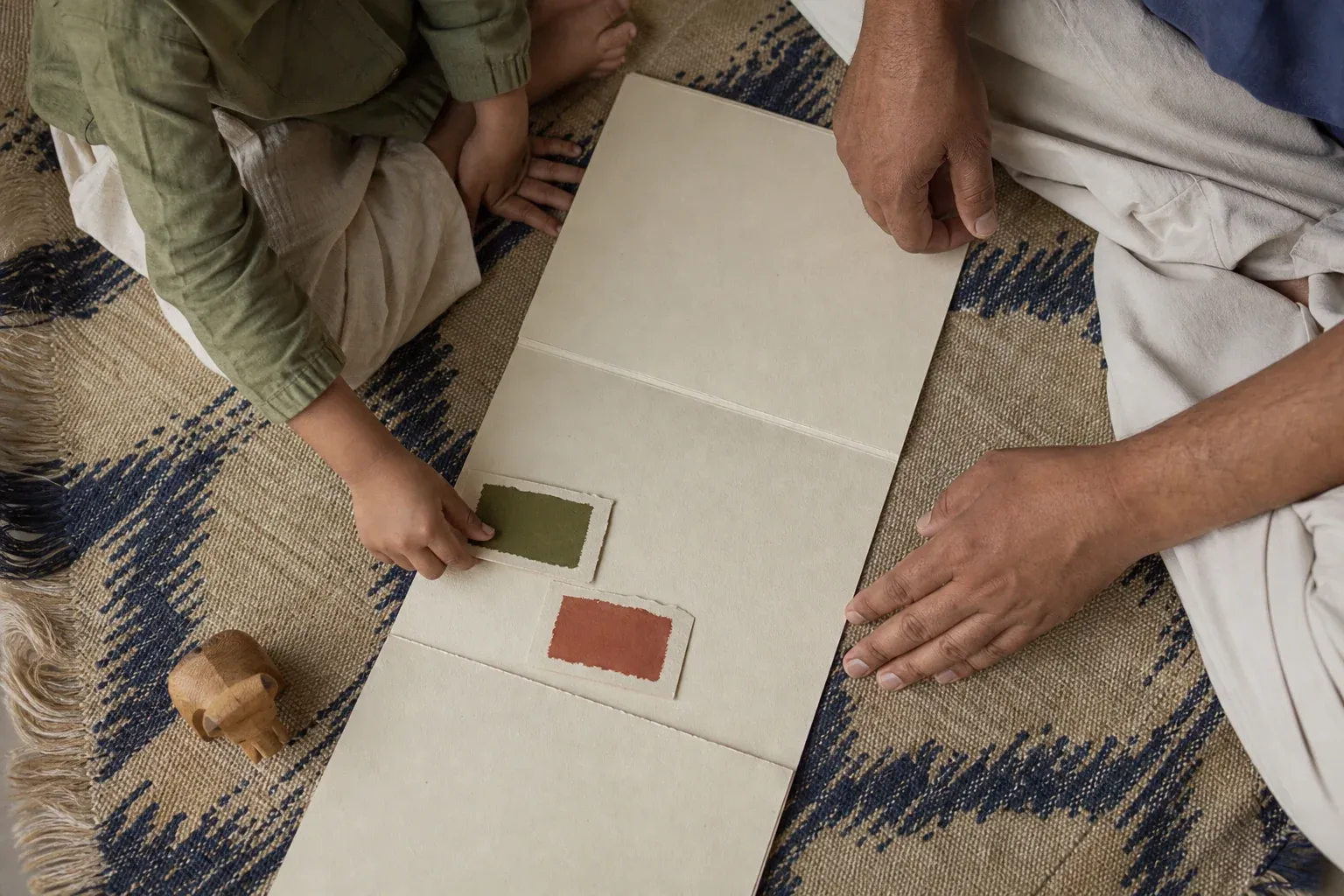 A child chooses between two pigment swatches on an unbound paper story dummy while an adult waits beside them.